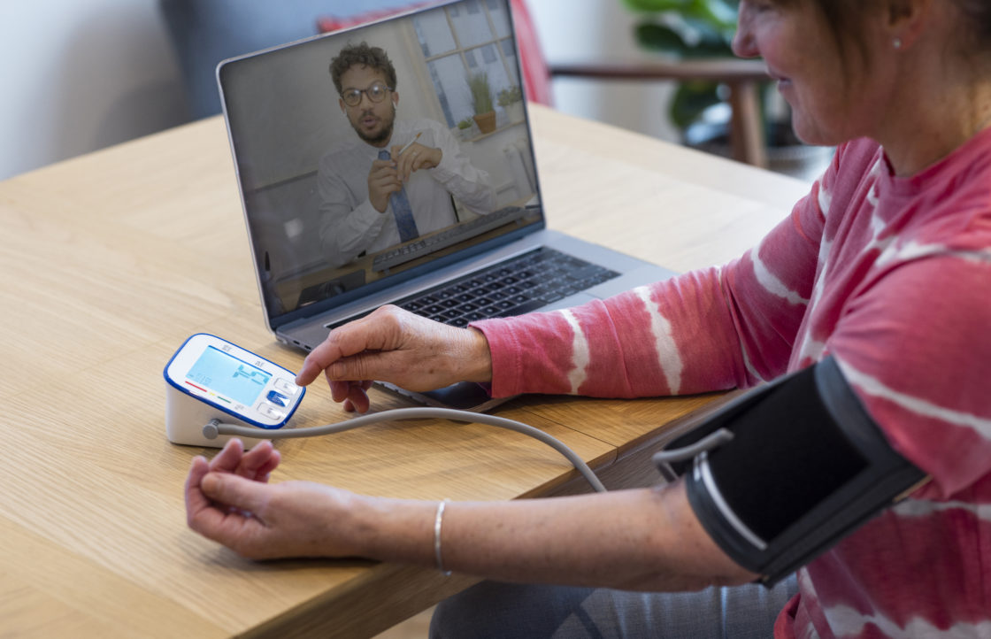 Woman in a virtual ward using a laptop to contact a medical professional about her health. She is siting at a table in a kitchen and is taking her blood pressure with a blood pressure gauge. She is in the North East of England.